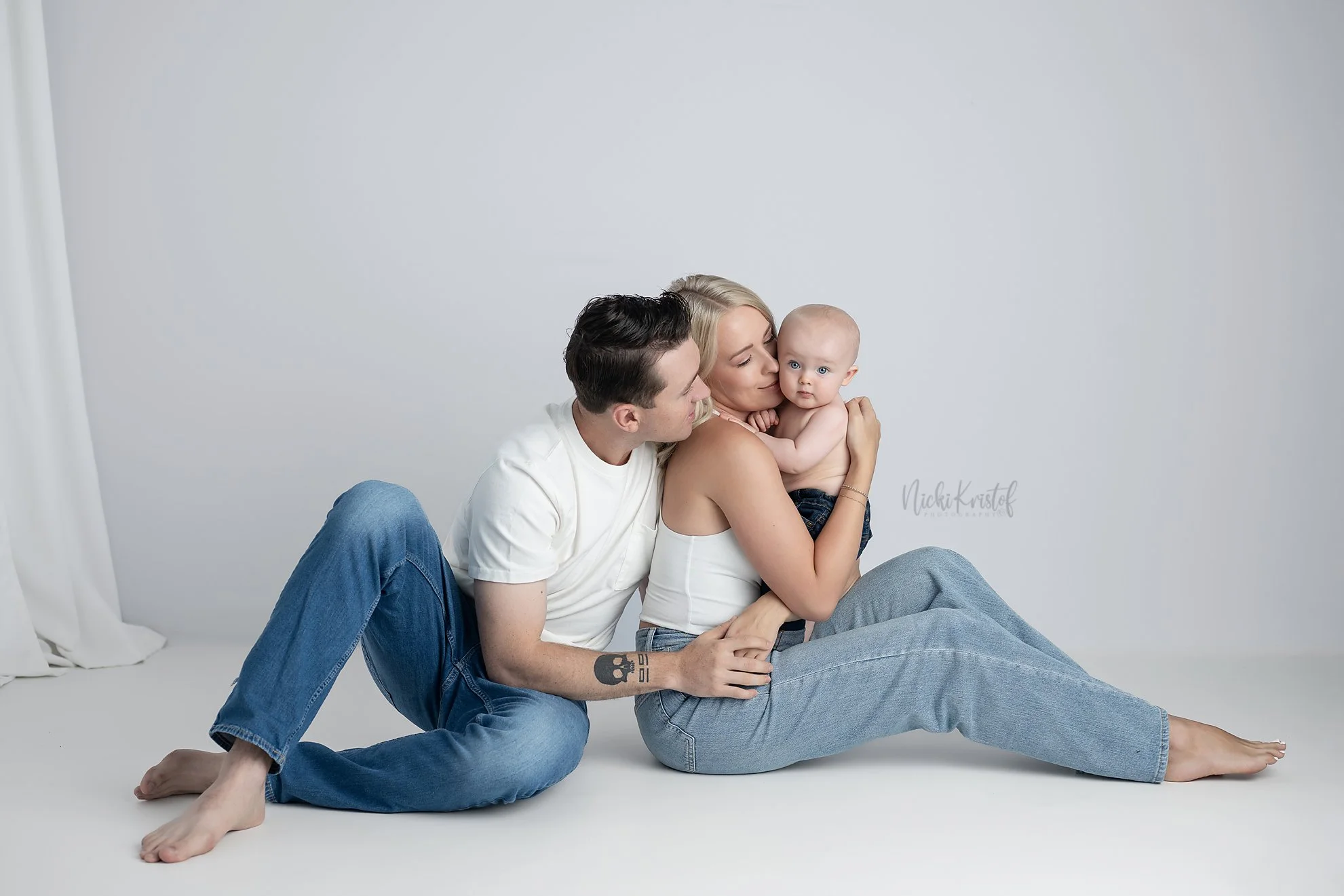 A family of three sitting on the floor against a plain white background. The man has dark hair and is wearing a white T-shirt and blue jeans. The woman has blonde hair, is wearing a white tank top and blue jeans, and is holding a baby with blue eyes 
