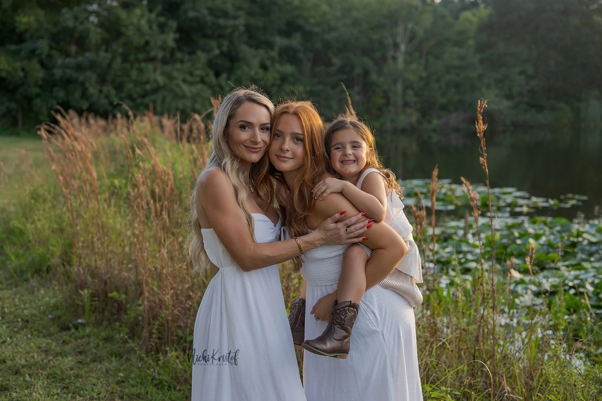 A woman and two young girls smiling and hugging near a lakeside with tall grass and trees in the background.