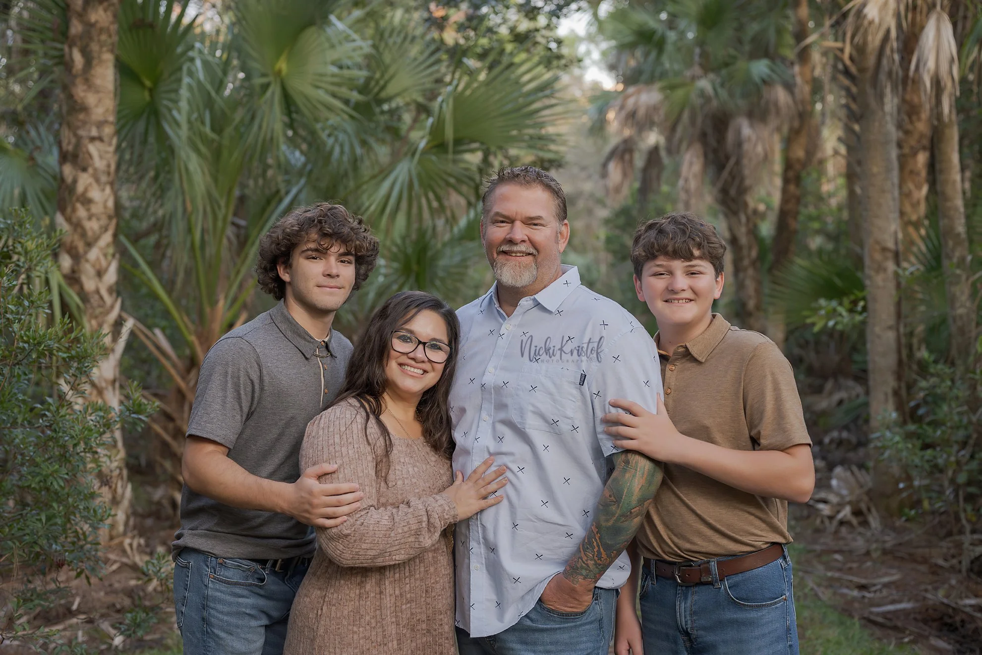 A smiling family of five standing outdoors among tropical trees, with their arms around each other.