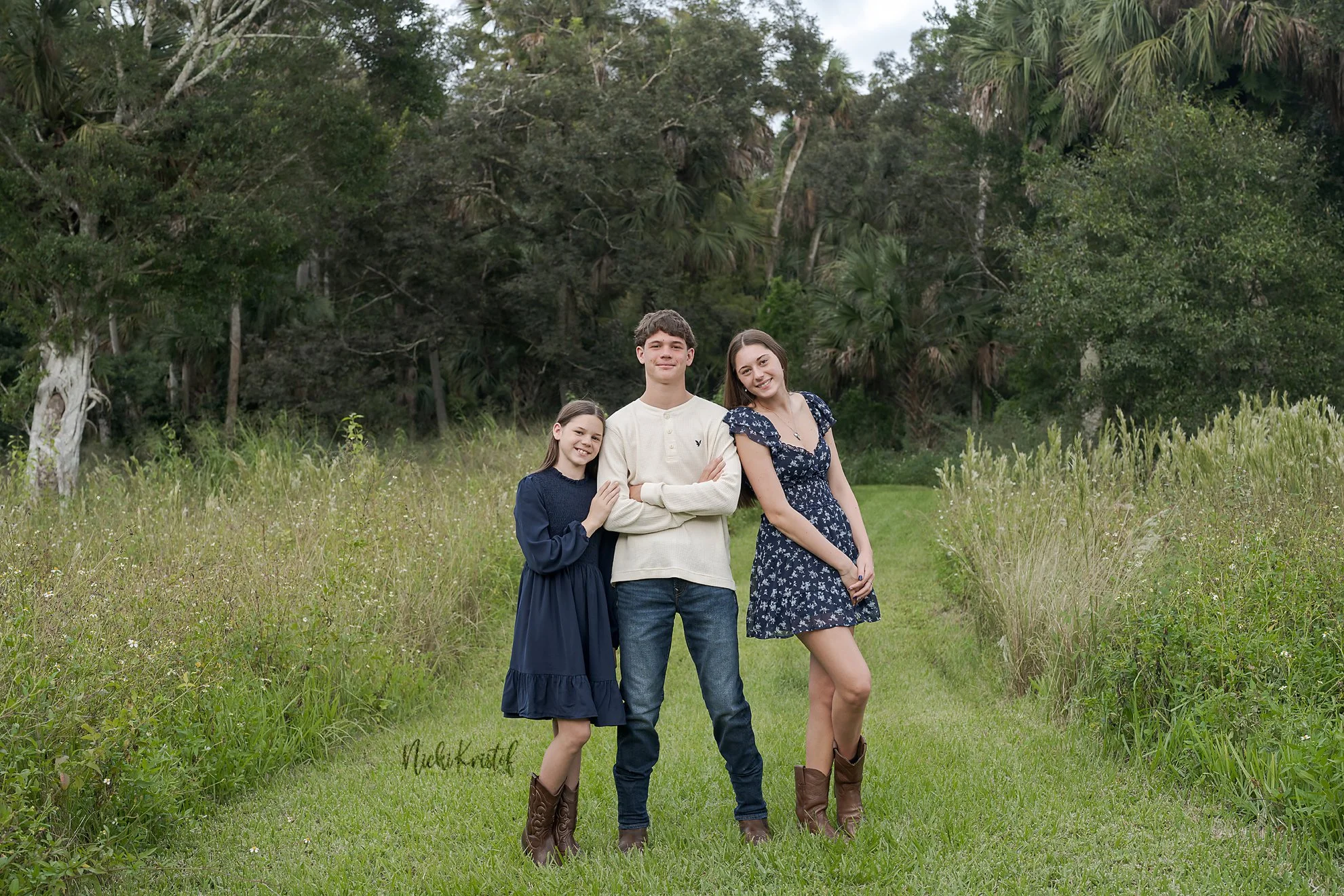 Four teenagers, two boys and two girls, standing on a grassy path in front of a dense green forest, smiling at the camera.