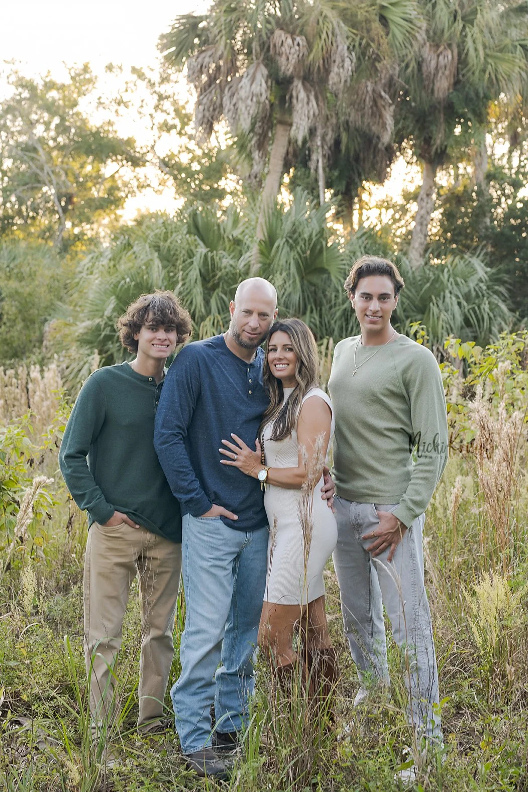 A family of four standing outdoors in a natural setting with trees and tall grass, smiling at the camera, during late afternoon or early evening