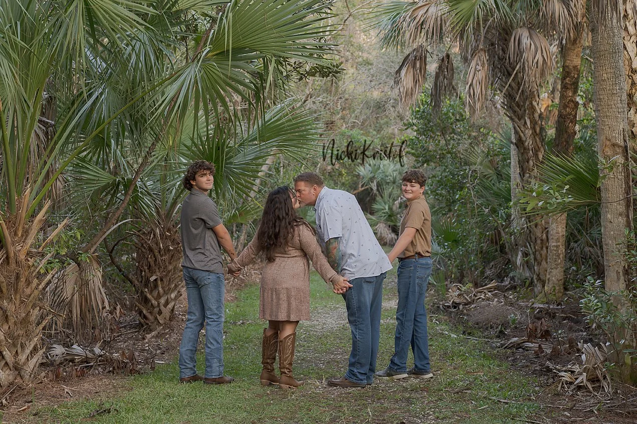 A family of five holding hands in a circle outdoors surrounded by large green palm trees, with one person kissing another on the lips and another person smiling.