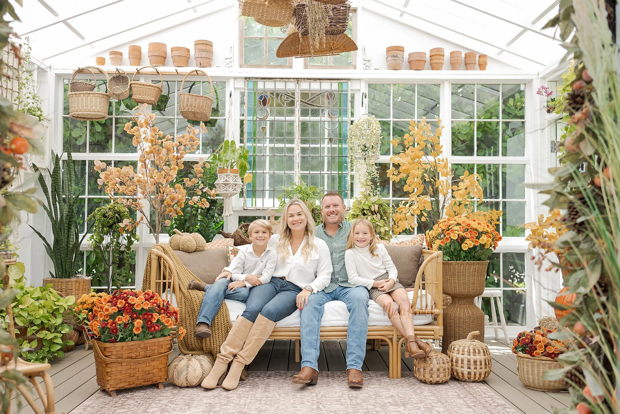 A family of five, including a mother, father, and three children, sitting together on a sofa inside a sunlit greenhouse filled with various potted plants and orange flowers.
