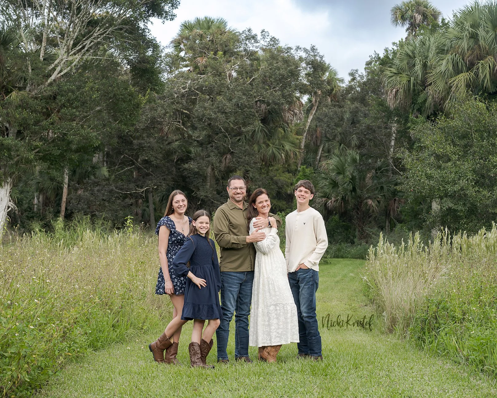 Family of five standing on a grassy field with trees in the background, smiling for a photo