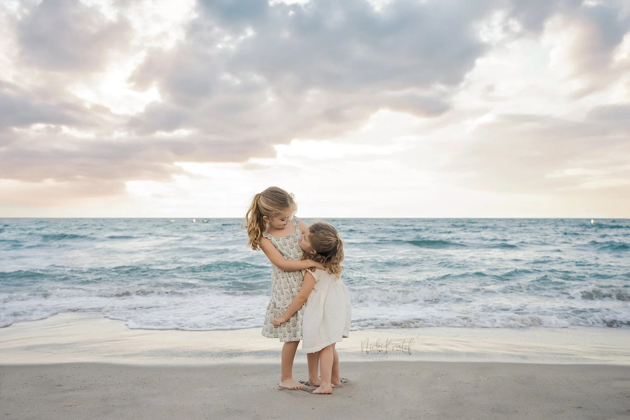 Two young girls embracing on the beach with the ocean and cloudy sky in the background.