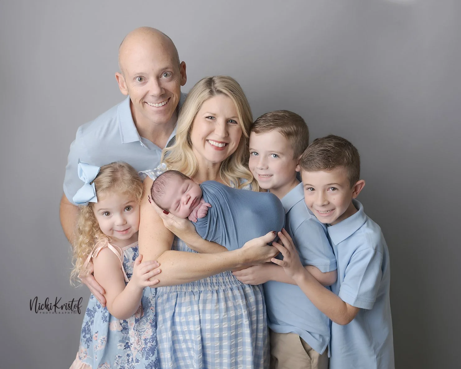 Family of seven poses against a plain gray background, with a woman holding a newborn wrapped in blue cloth, and four children around her, one girl with a blue bow and boy and girls smiling.