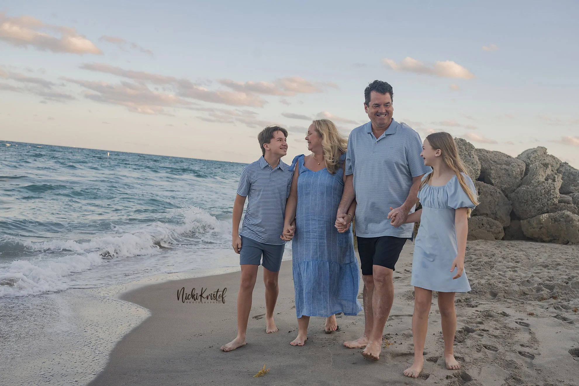 A family of four holding hands and walking on the beach near rocky shoreline during sunset. The family consists of a woman with blonde hair, a man with dark hair, a teenage boy, and a young girl, all wearing casual summer clothes.