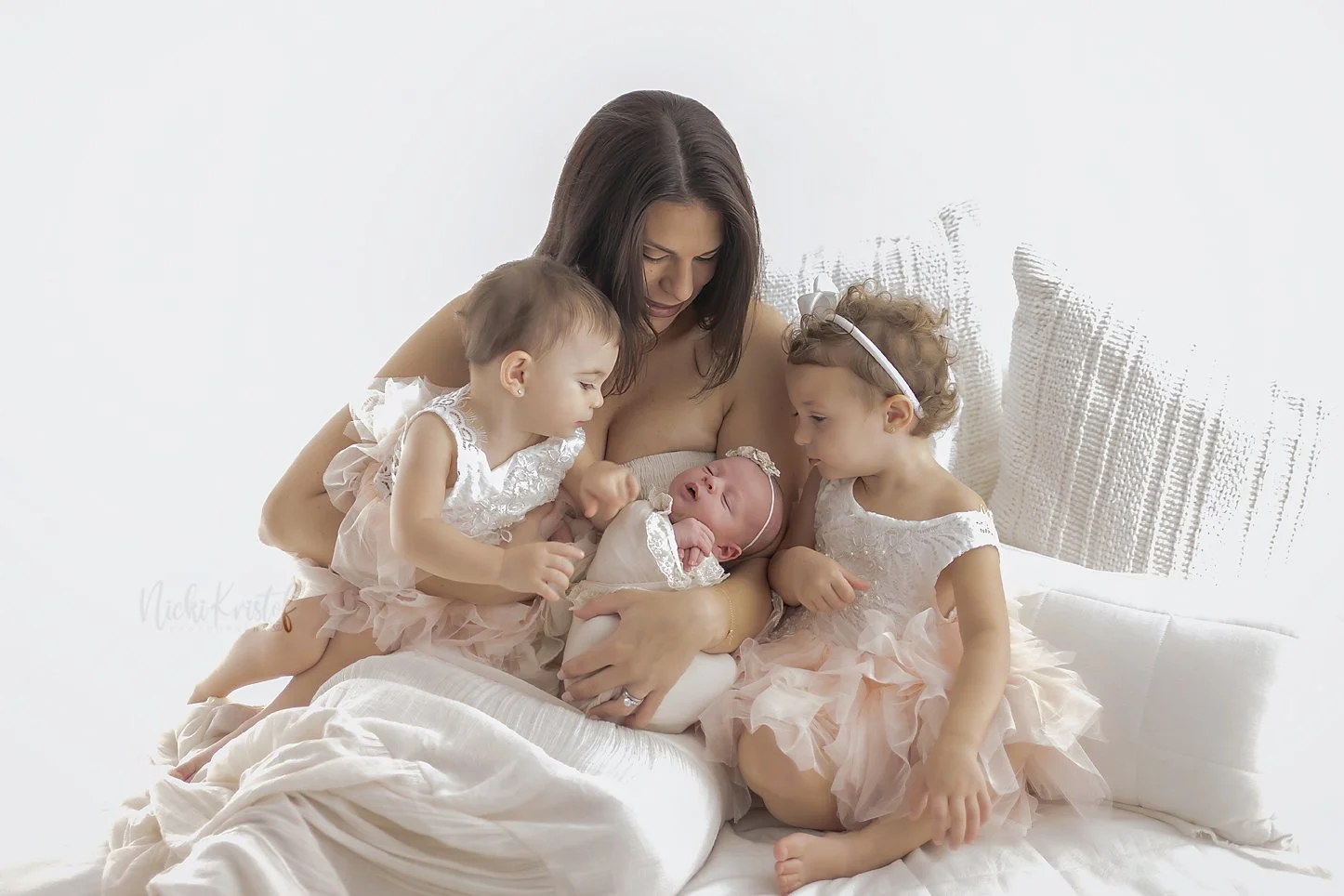 A mother holding her newborn baby on her lap while two young girls, wearing matching dresses, are leaning in to see the baby, all sitting on a white bed. newborn lifestyle family portrait.