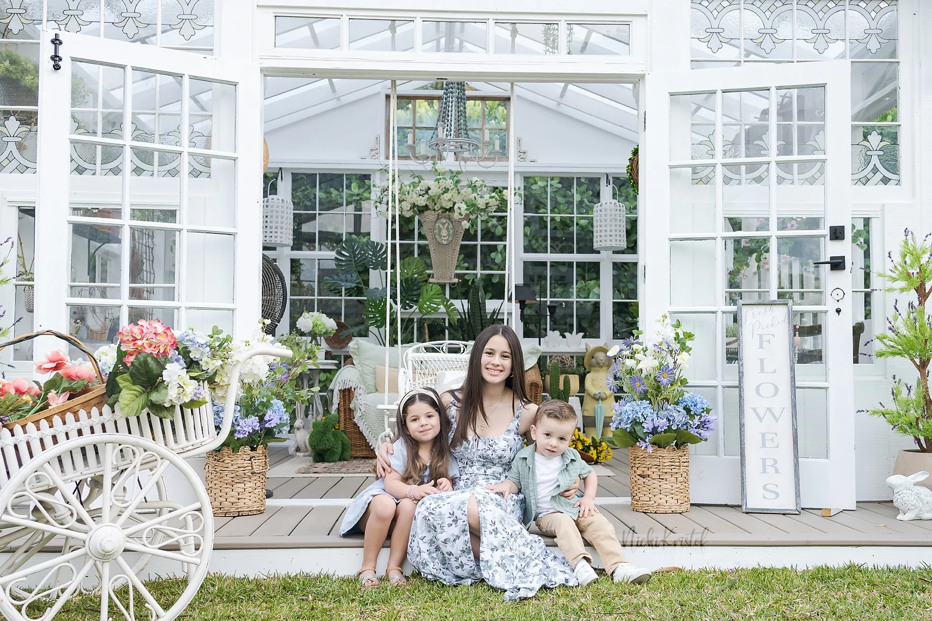 A woman and three children sitting in front of a white garden shed decorated with flowers and plants, with a sign reading "Flower" on the right side.