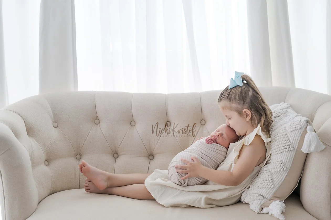 Young girl holding a newborn baby on a beige sofa in front of white curtains.