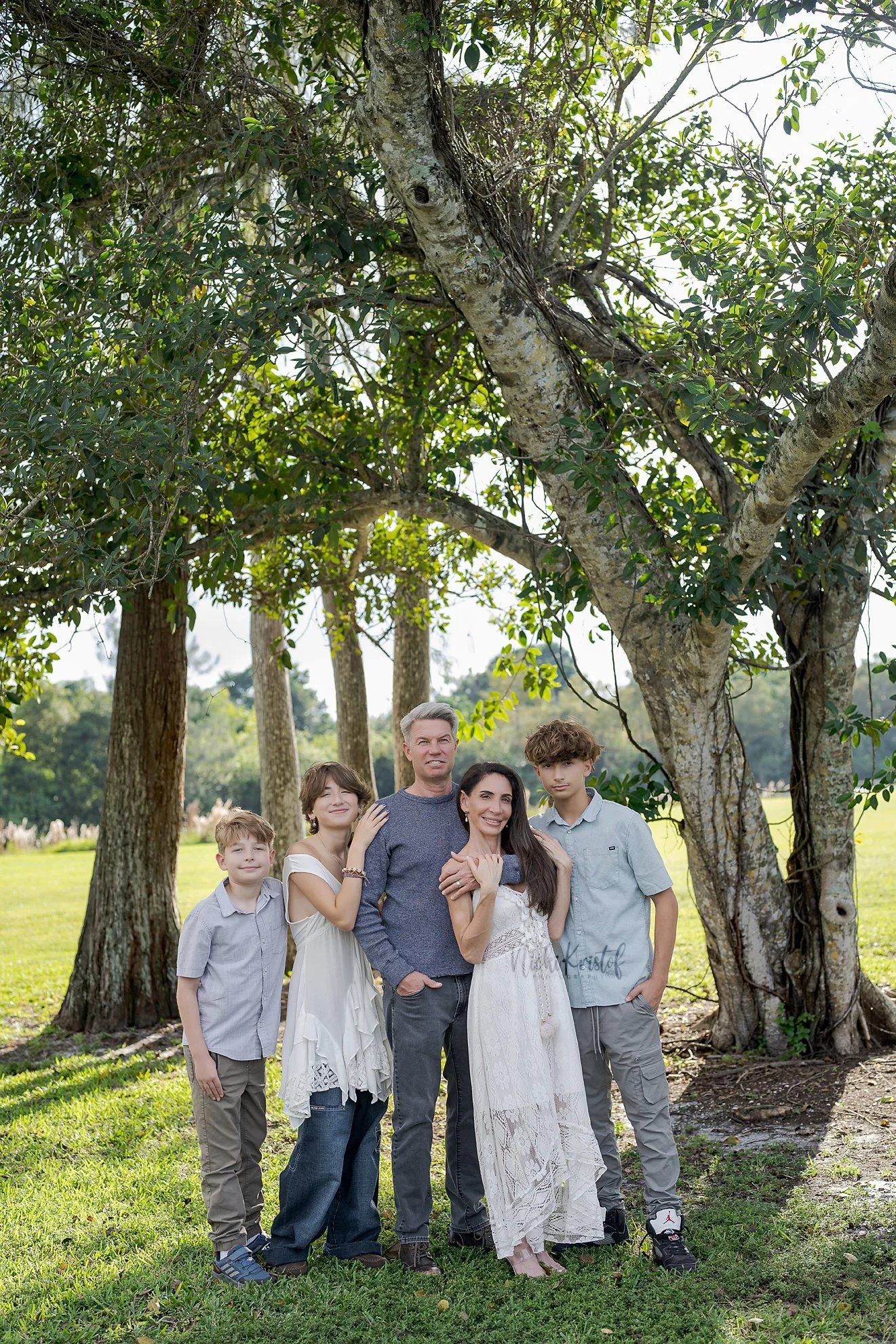 A family of six people standing outdoors in front of trees on a sunny day, smiling and posing for the photo.
