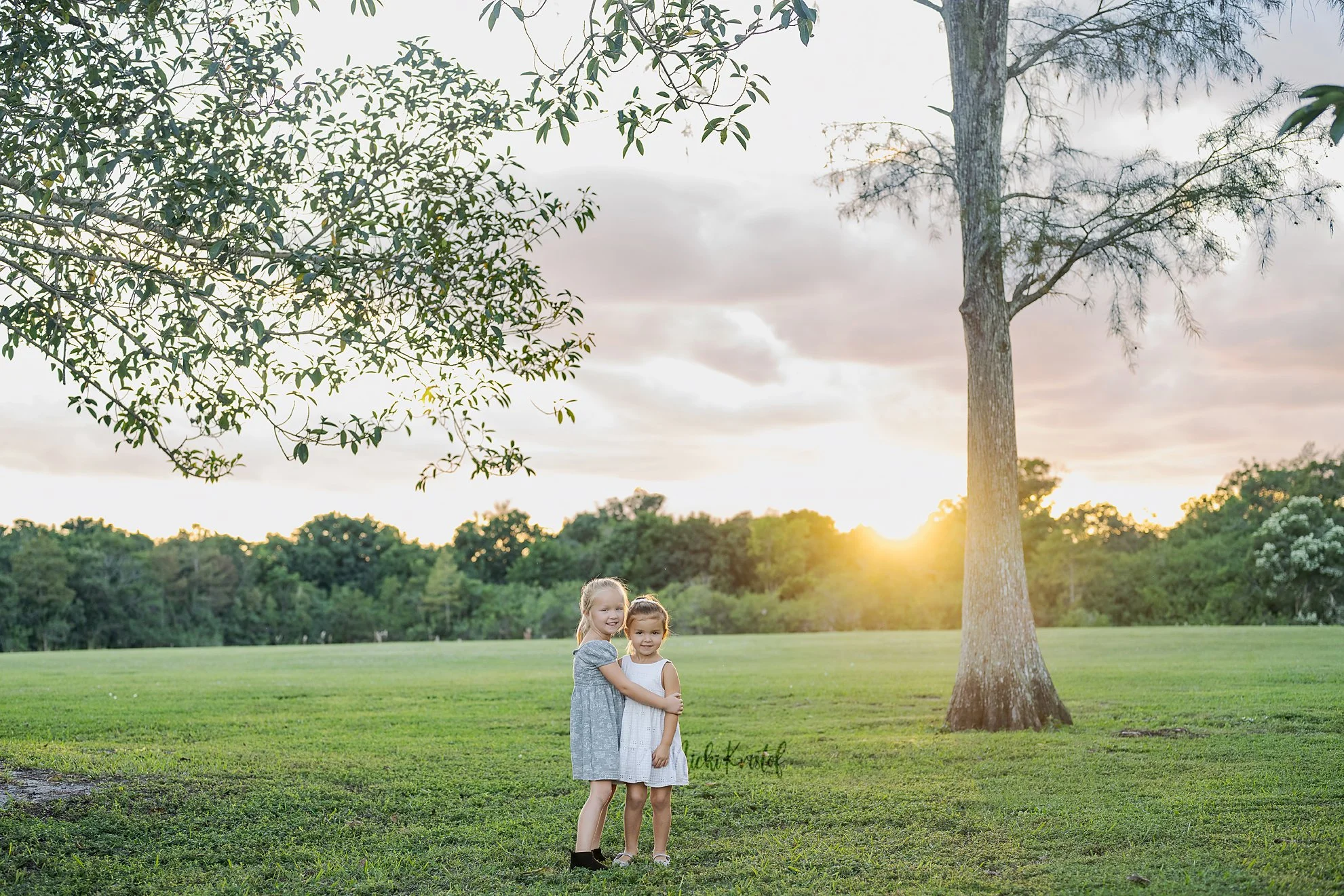 Two young girls standing outdoors on the grass in a park at sunset, smiling and hugging each other with trees in the background.