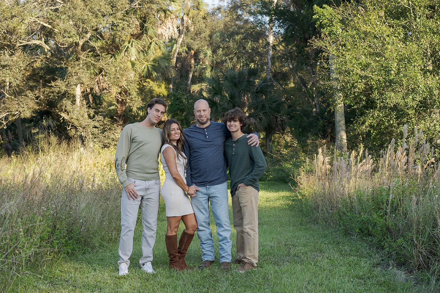 A family of four posing outdoors on a grassy path with trees and bushes in the background.