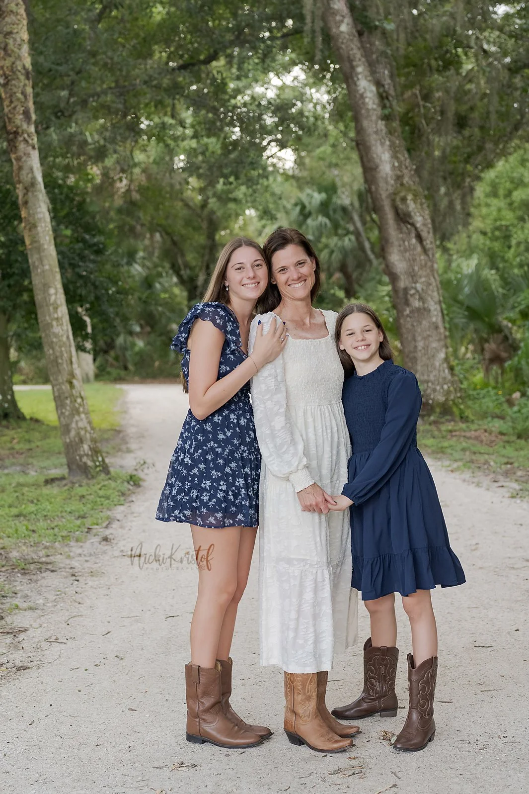 Three women smiling and holding hands outdoors on a dirt path surrounded by trees.
