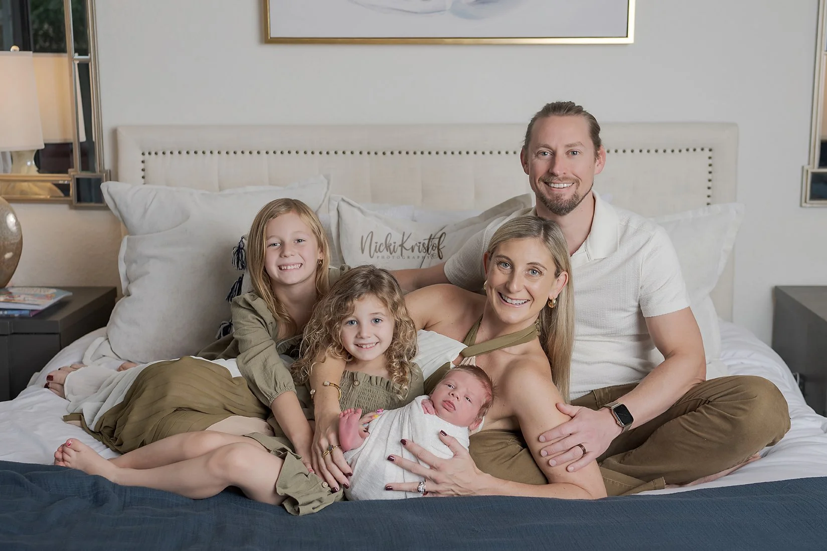 A family of six, including two adults and four children, sitting on a bed in a bedroom and smiling at the camera. newborn lifestyle family portrait.