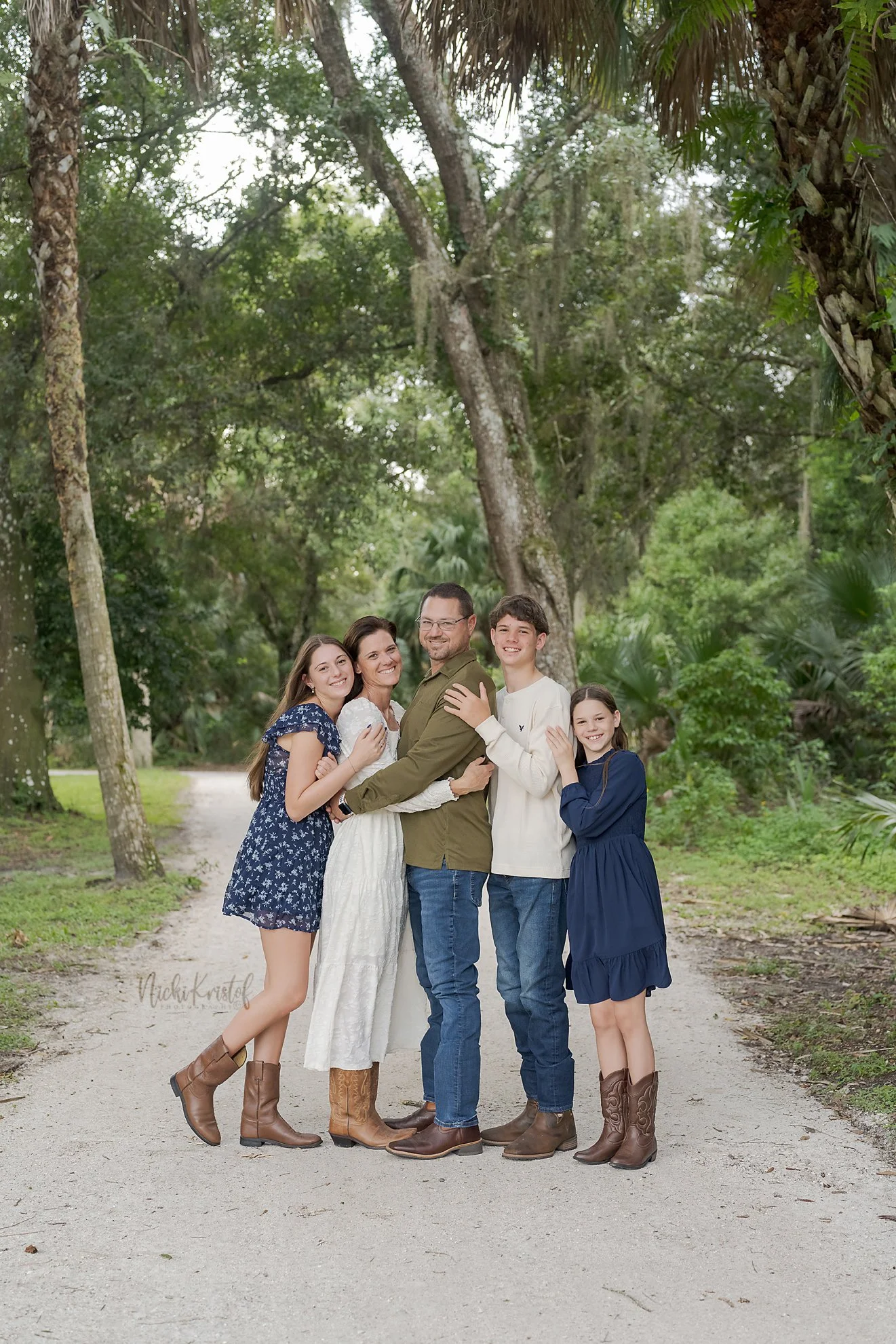 Family of five standing on a dirt path in a forest, smiling and embracing each other. The family includes a woman in a white dress, a man in a green shirt, and three children, two girls and one boy, all wearing casual outfits and cowboy boots.