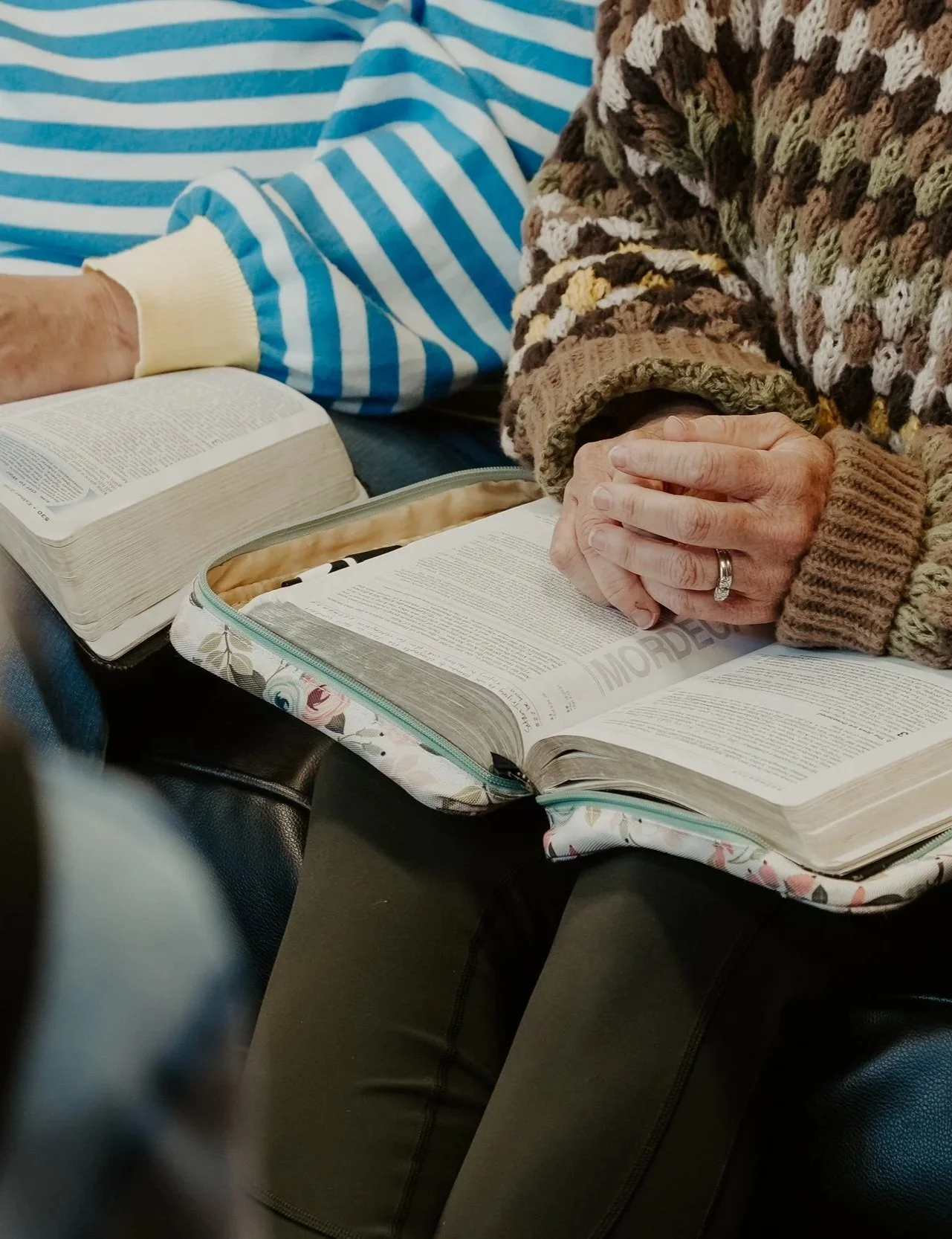 Close-up of elderly person's hands clasped together over an open Bible, with another Bible and a patterned bag nearby, and a person wearing a multicolored sweater and a striped blue and white shirt sitting beside.