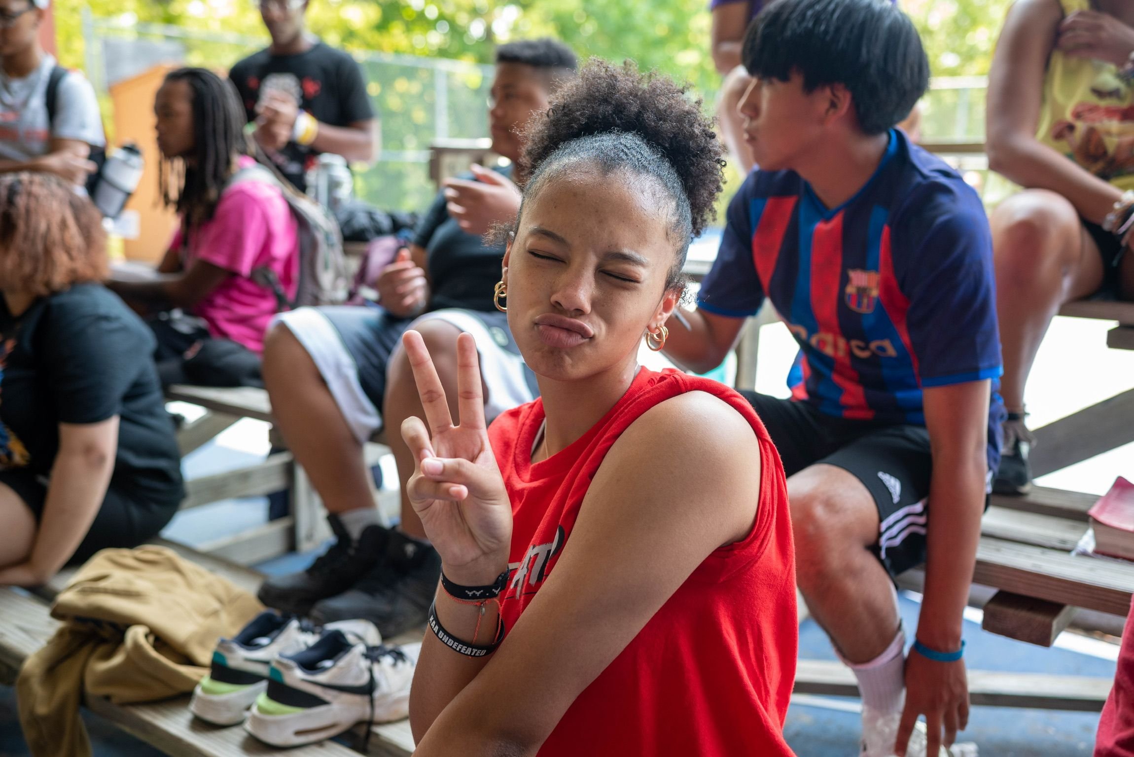 A young girl in a red sleeveless shirt making a peace sign with her fingers, with her eyes closed and lips puckered, sitting among a group of people outdoors during daytime, surrounded by trees and benches.