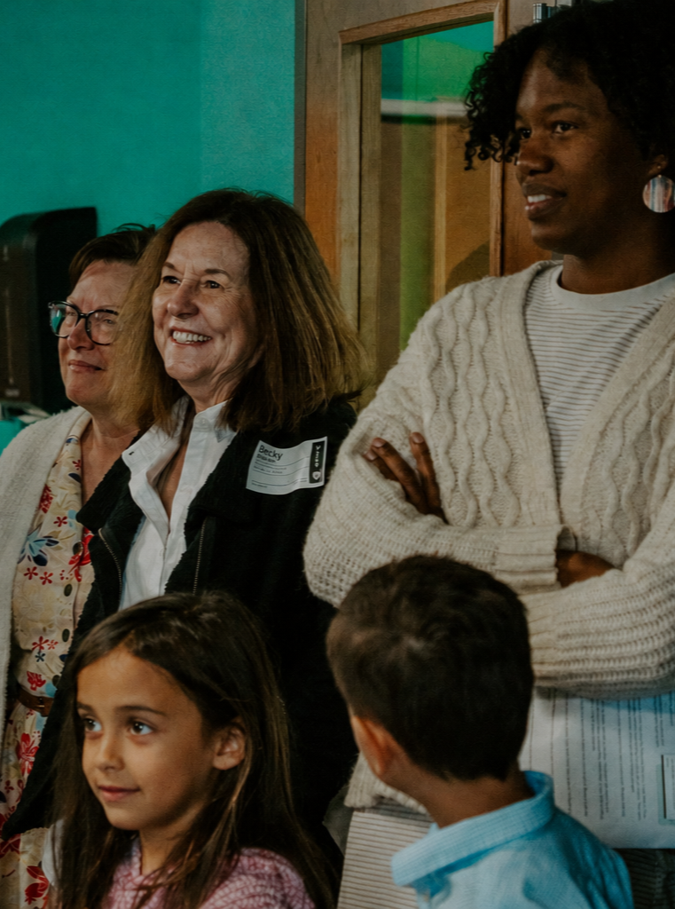 A diverse group of people, including children and adults, gathered together indoors, smiling and listening attentively.