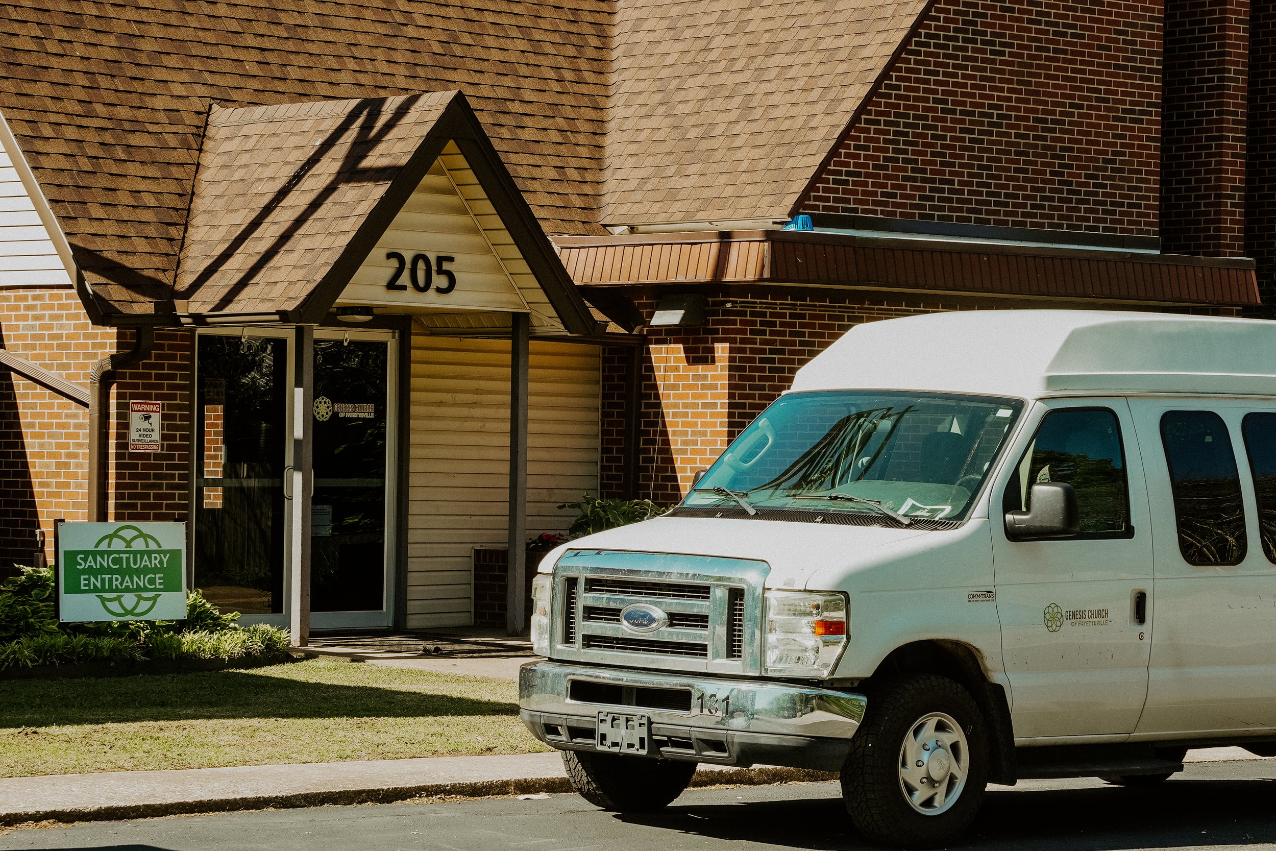 A white van parked in front of the Genesis Church of Fayetteville. The church building has brick and siding exterior with an entrance marked by a green sign that reads 'Sanctuary Entrance.' The building has a prominent triangular roof over the entrance. Shadows of utility poles are cast across the roof.