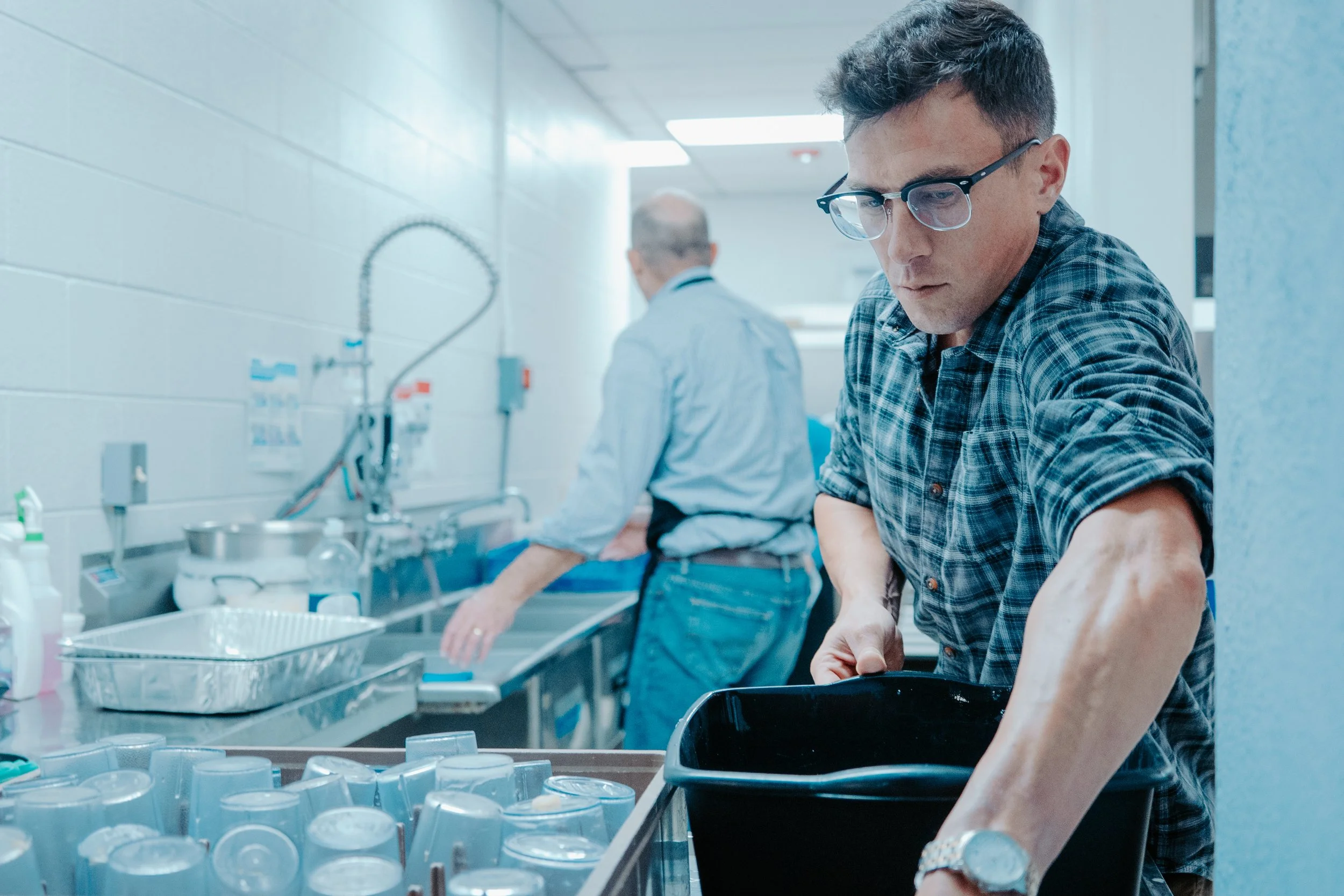 Two people working in a commercial kitchen, washing dishes at a stainless steel sink.
