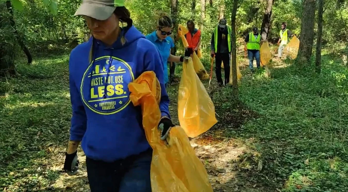 Group of volunteers participating in a cleanup event in a forest, wearing gloves and some holding yellow trash bags, collecting litter along a wooded trail.