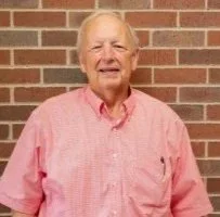 An elderly man with white hair and a friendly smile, wearing a pink shirt, standing in front of a brick wall.