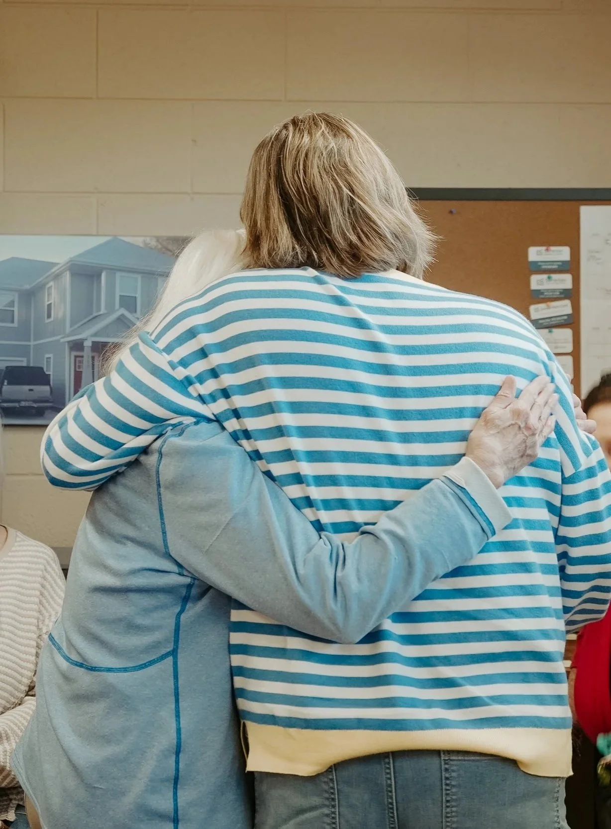 Two women hugging each other, one with blonde hair and a striped shirt, and the other with grey hair, in a room with a picture of a house on the wall.