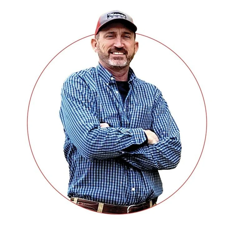 A man with a beard and mustache wearing a baseball cap and a blue checkered button-up shirt, standing with his arms crossed and smiling.