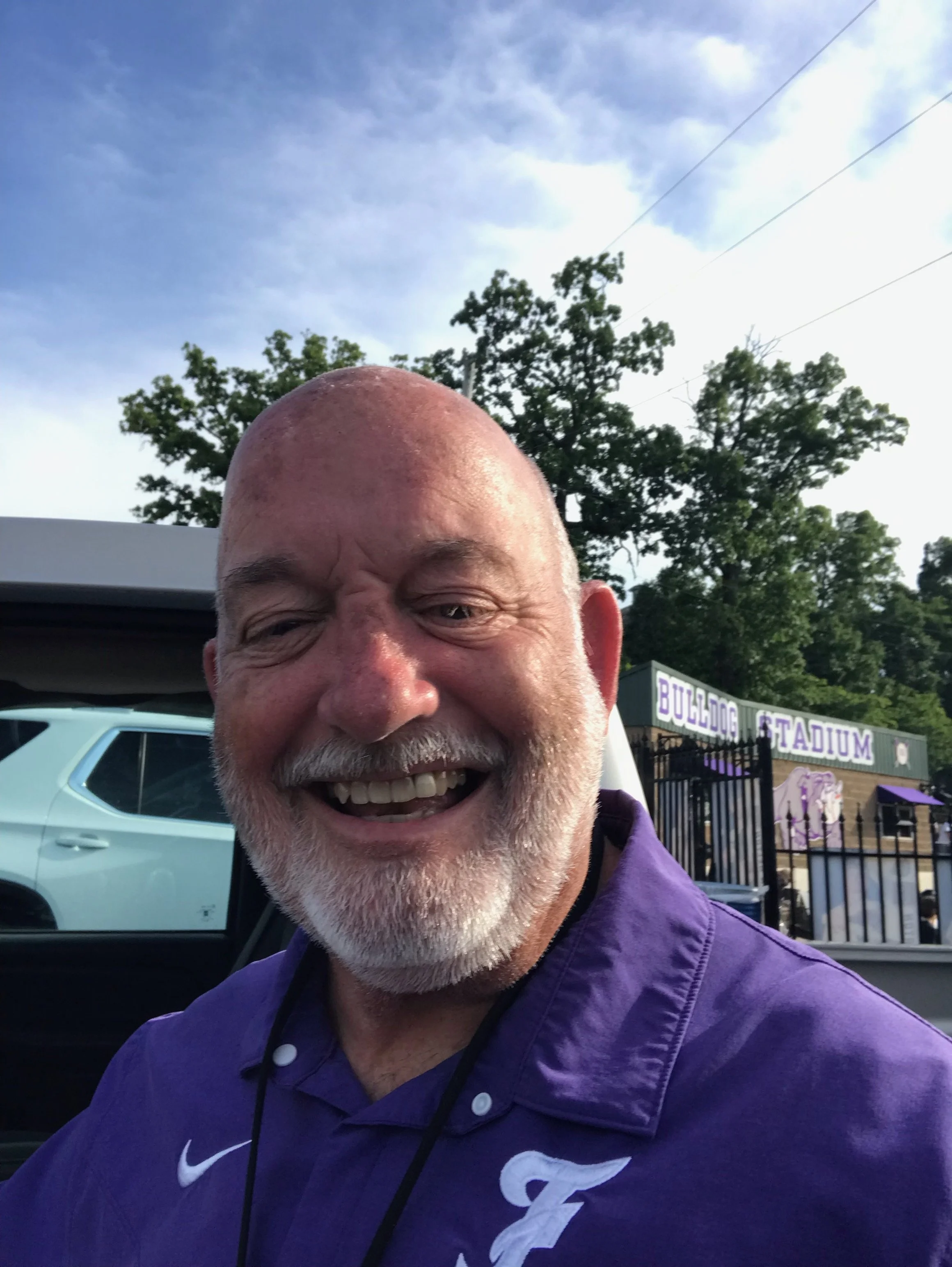 A smiling bald man with a beard, wearing a purple shirt with a white Nike logo and a 'G' letter. He is outdoors, standing in front of a light blue SUV, with trees and a sign for a stadium in the background.
