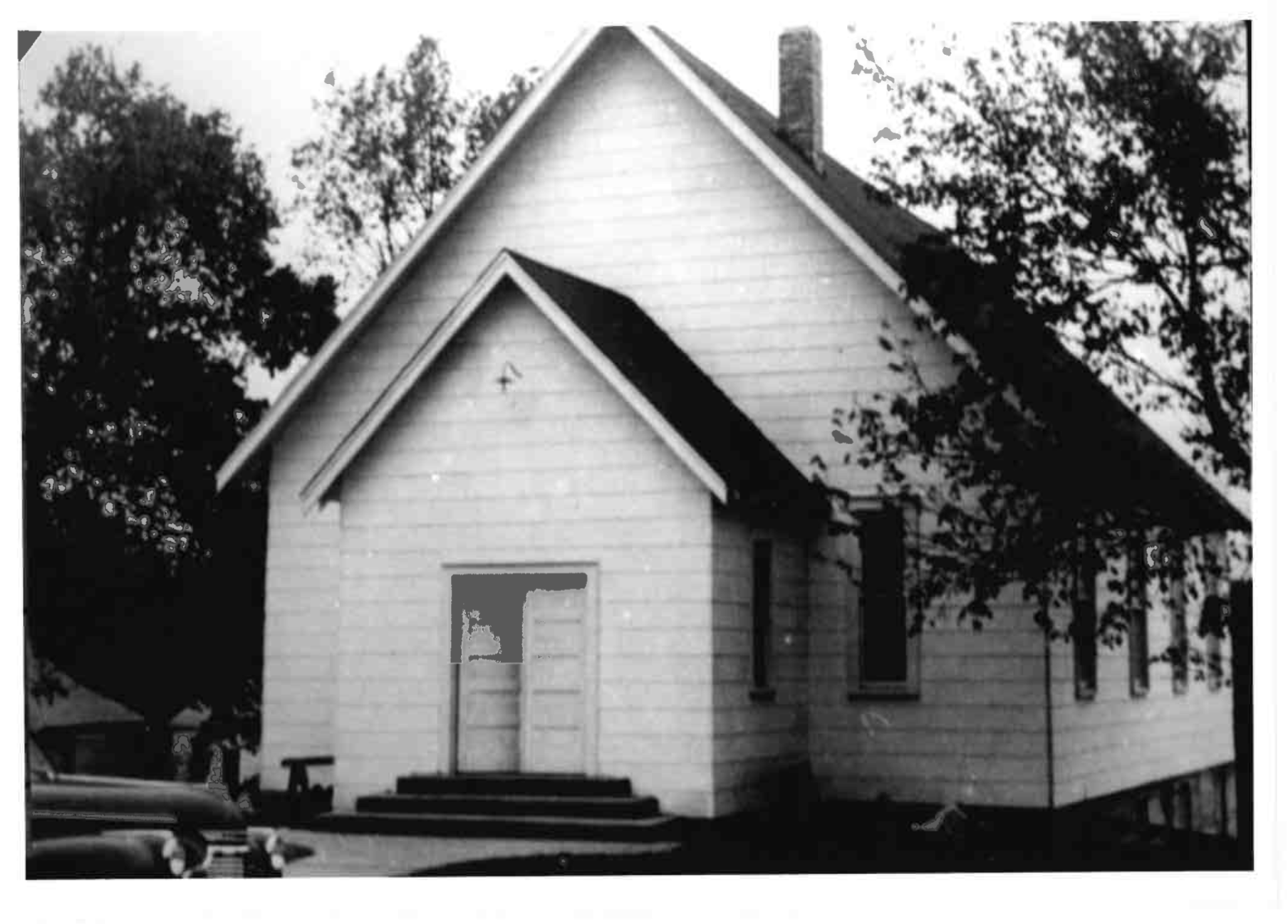 A black and white photograph of a house with steep gabled roof and horizontal siding, surrounded by trees.