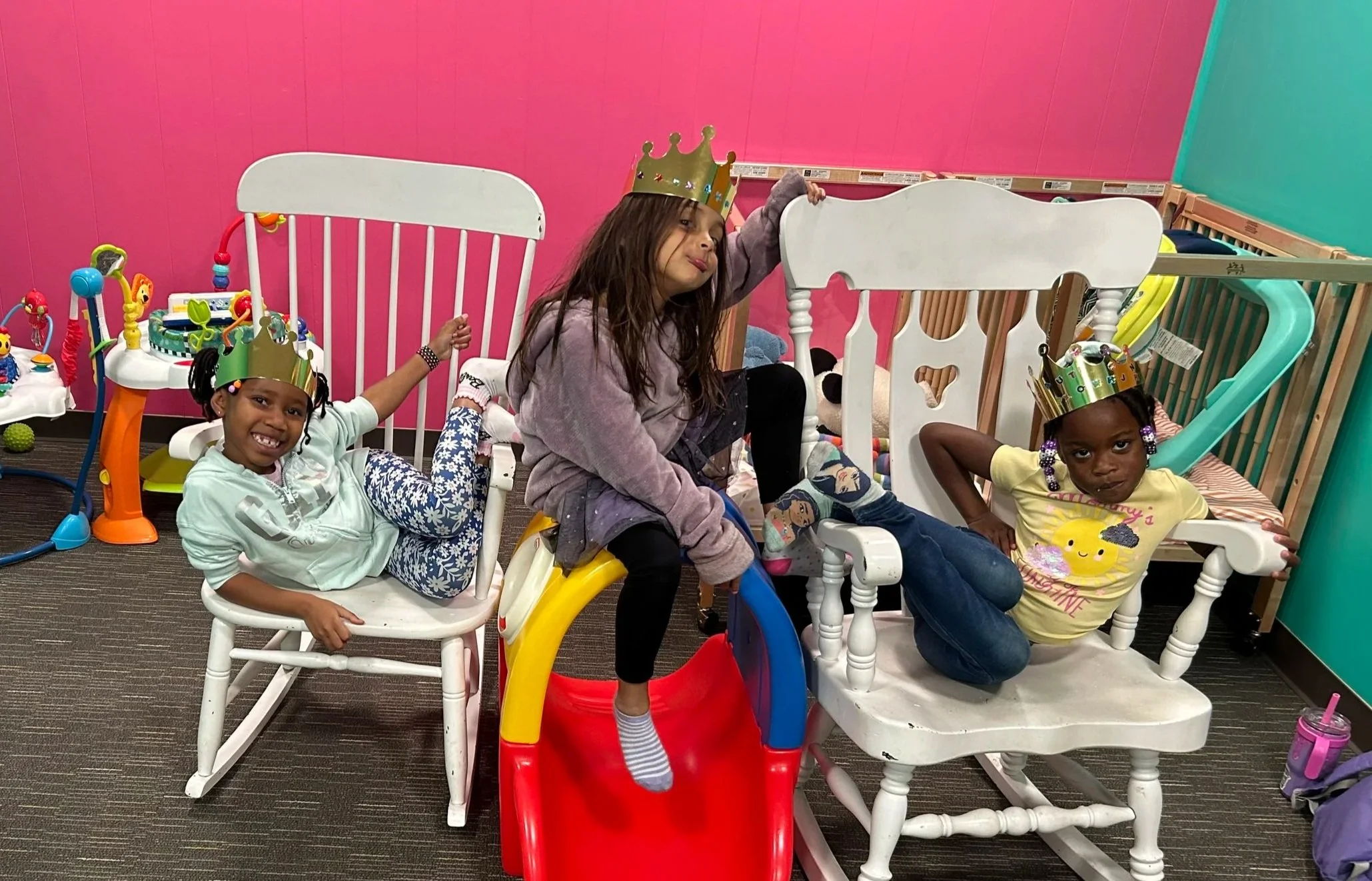 Children sitting and playing inside a colorful indoor play area, wearing paper crowns, with playground equipment and toys around them.