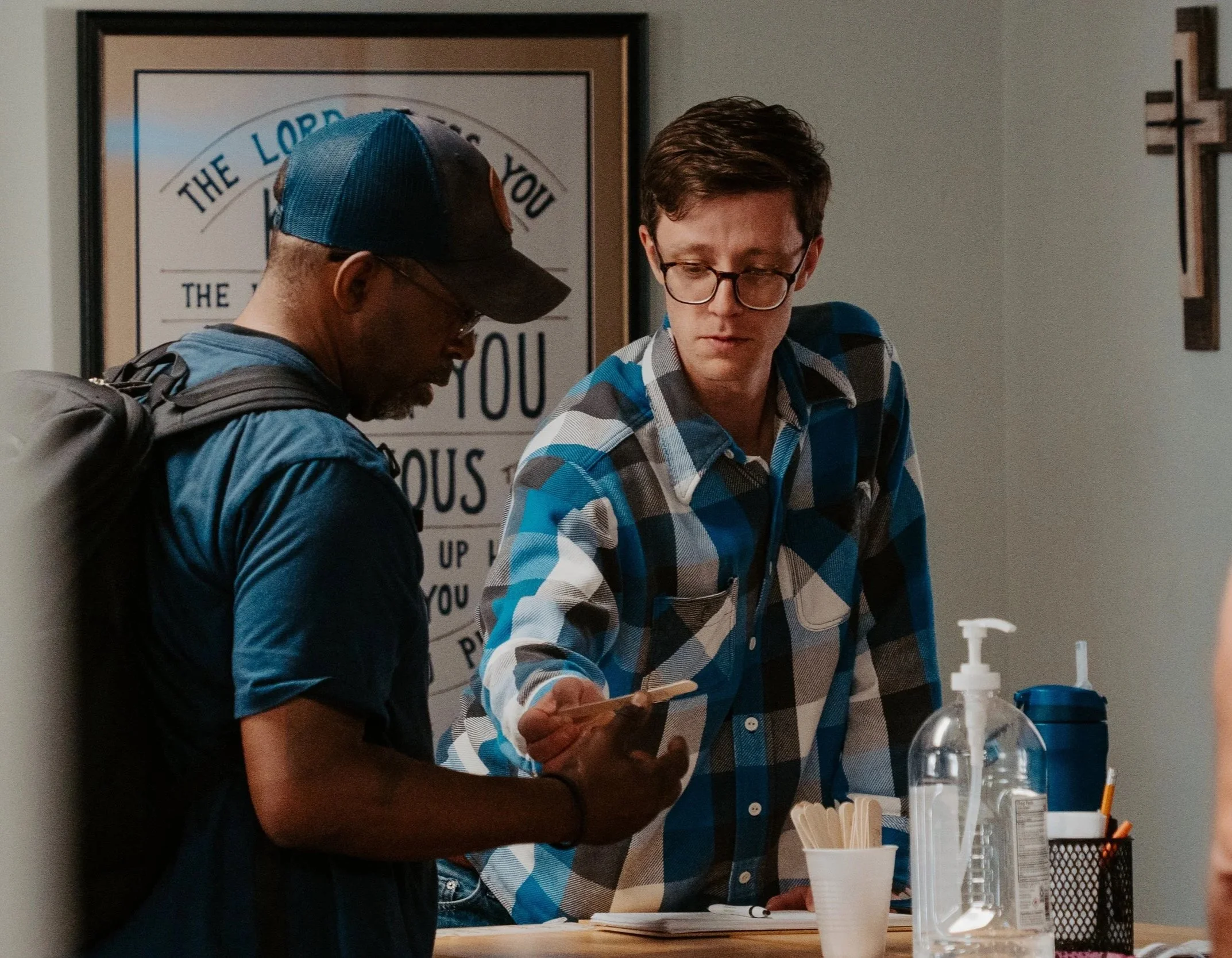 Two young men standing near a table with bottles of hand sanitizer, cups with wooden stir sticks, and a container of pens, in a room with a religious cross and framed sign on the walls.
