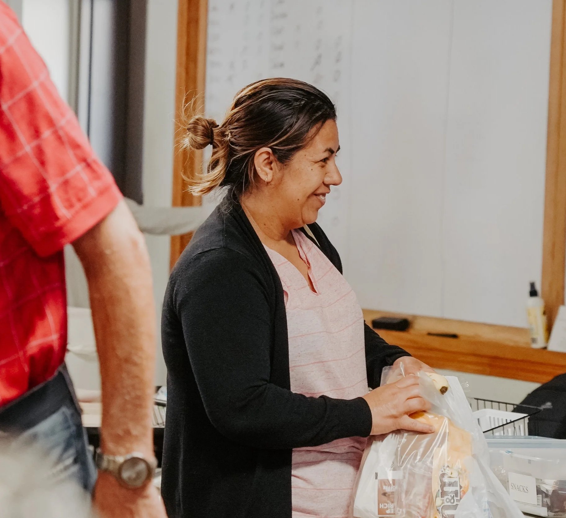 A woman with brown hair tied in a bun, wearing a pink and gray striped shirt and a black cardigan, is standing at a counter packaging bread. She is smiling and appears to be in a kitchen or grocery store. Partially visible in the foreground is a person wearing a red checkered shirt, with a wristwatch on their left arm.