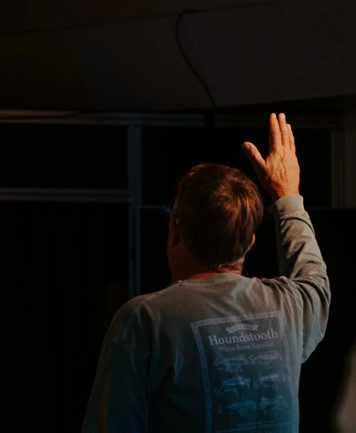 A person with short hair and glasses, wearing a gray shirt with 'Houndstooth' written on the back, is reaching up with their right hand towards the ceiling in a dark room.