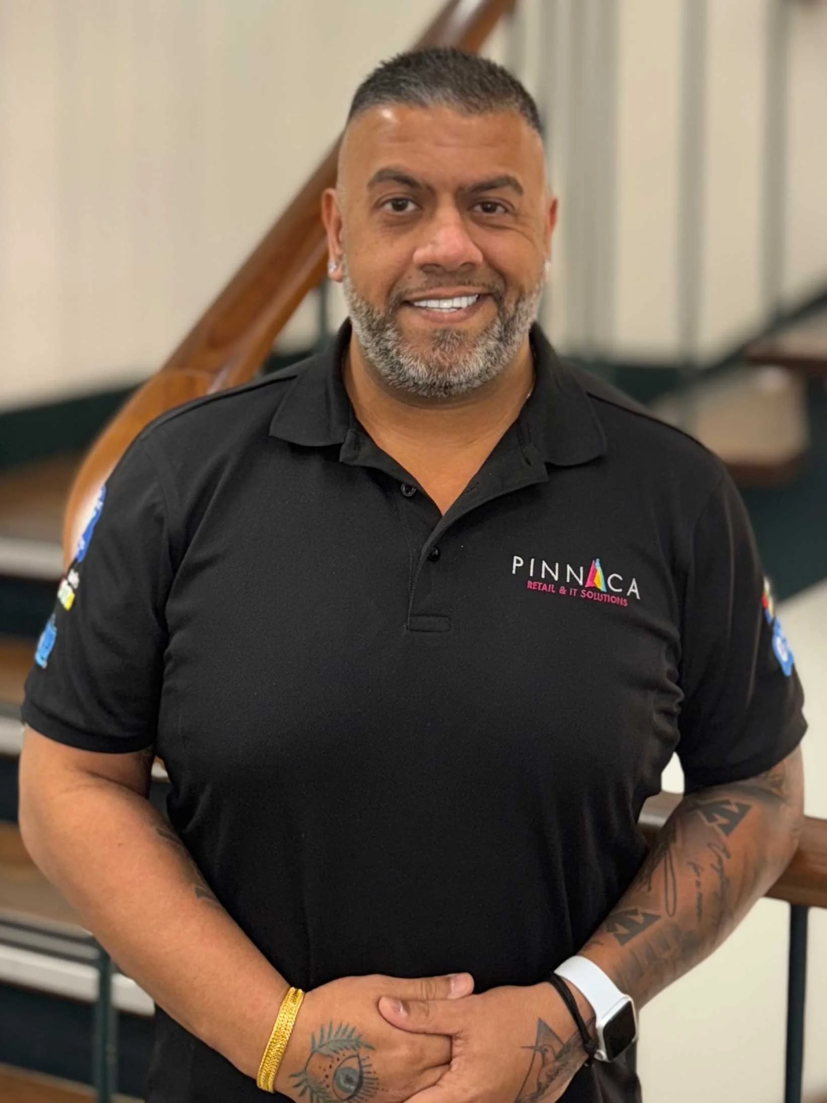 A smiling man with short hair and tattoos on his arms, wearing a black polo shirt with a logo that says Pinnaca Retail & IT Solutions, standing indoors near a staircase.