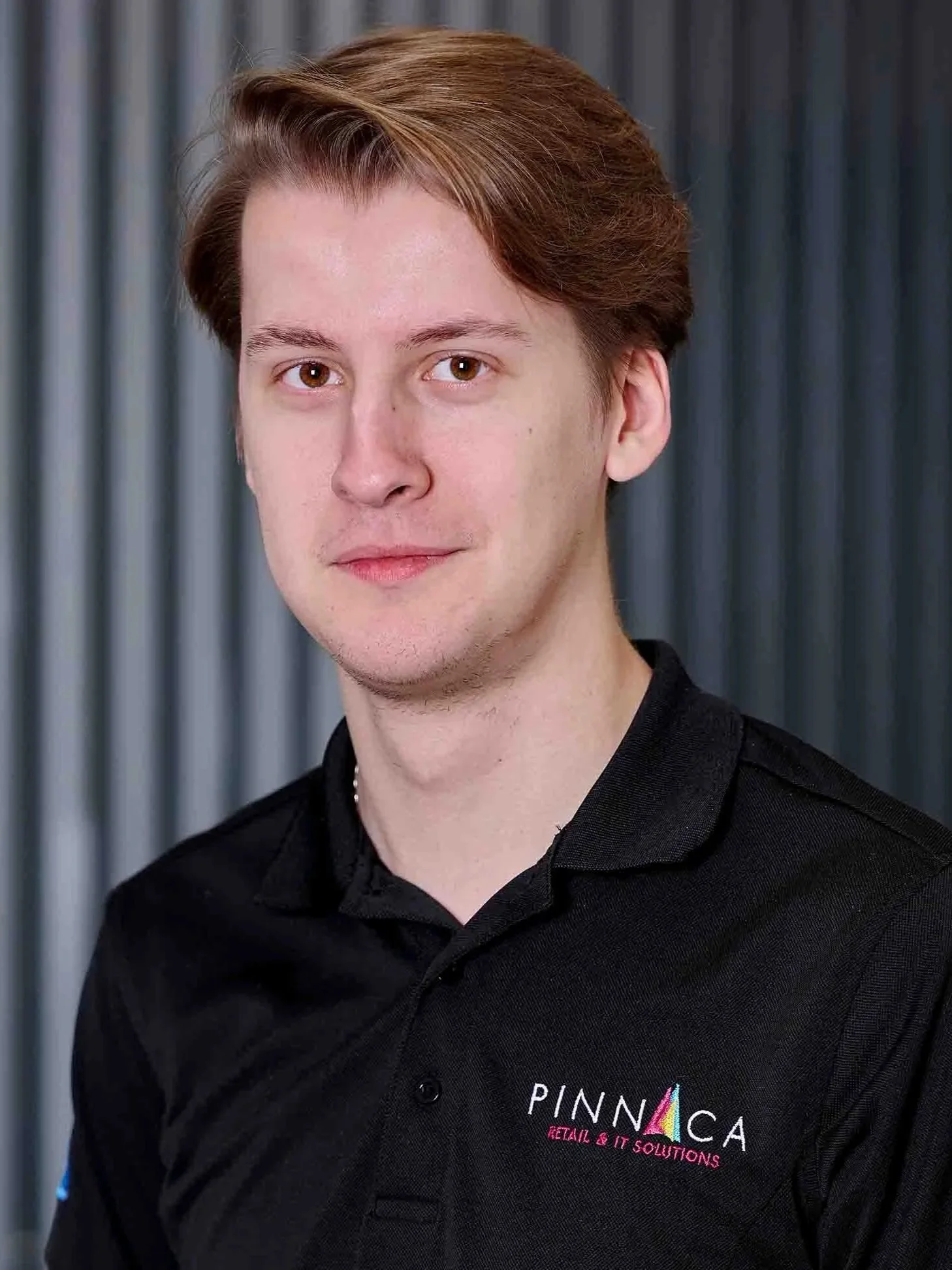Young man with light skin, light brown hair, wearing a black polo shirt with logos, standing in front of a grey striped background.
