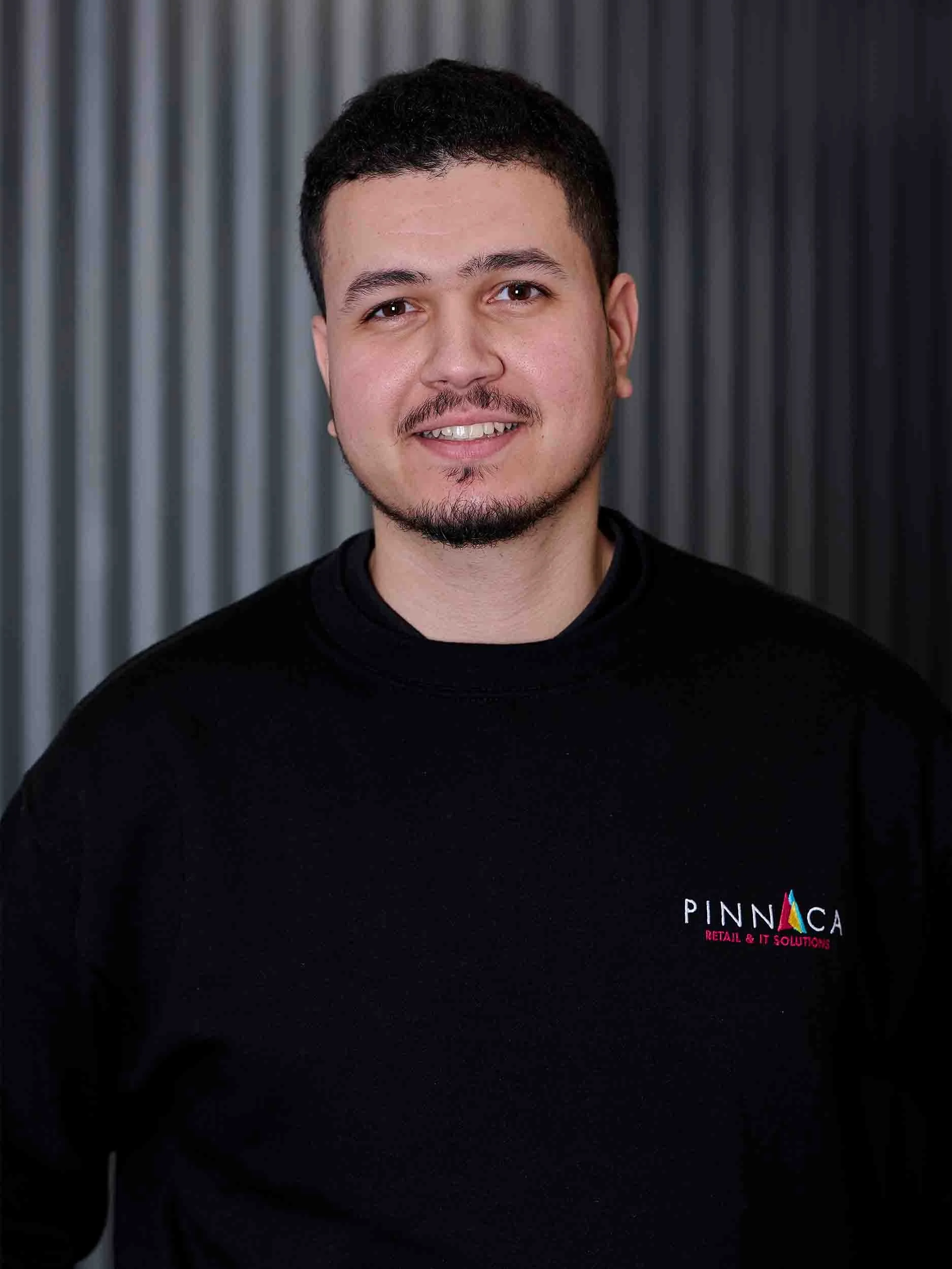 A young man with short dark hair, a goatee, and a slight smile, wearing a black sweatshirt with a colorful logo on the left chest that reads 'PINNICA RETAIL & IT SOLUTIONS', standing in front of a background with vertical dark gray and black stripes.
