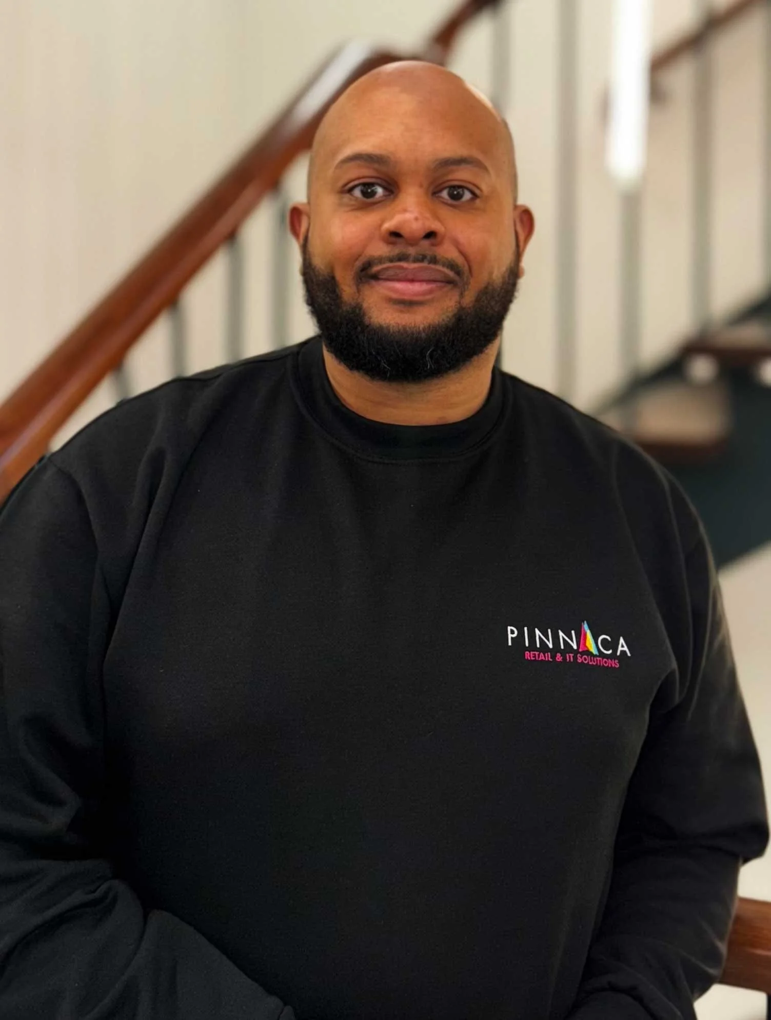 A man with a beard and bald head wearing a black sweatshirt with a logo that reads PinnovA, standing indoors near a staircase.