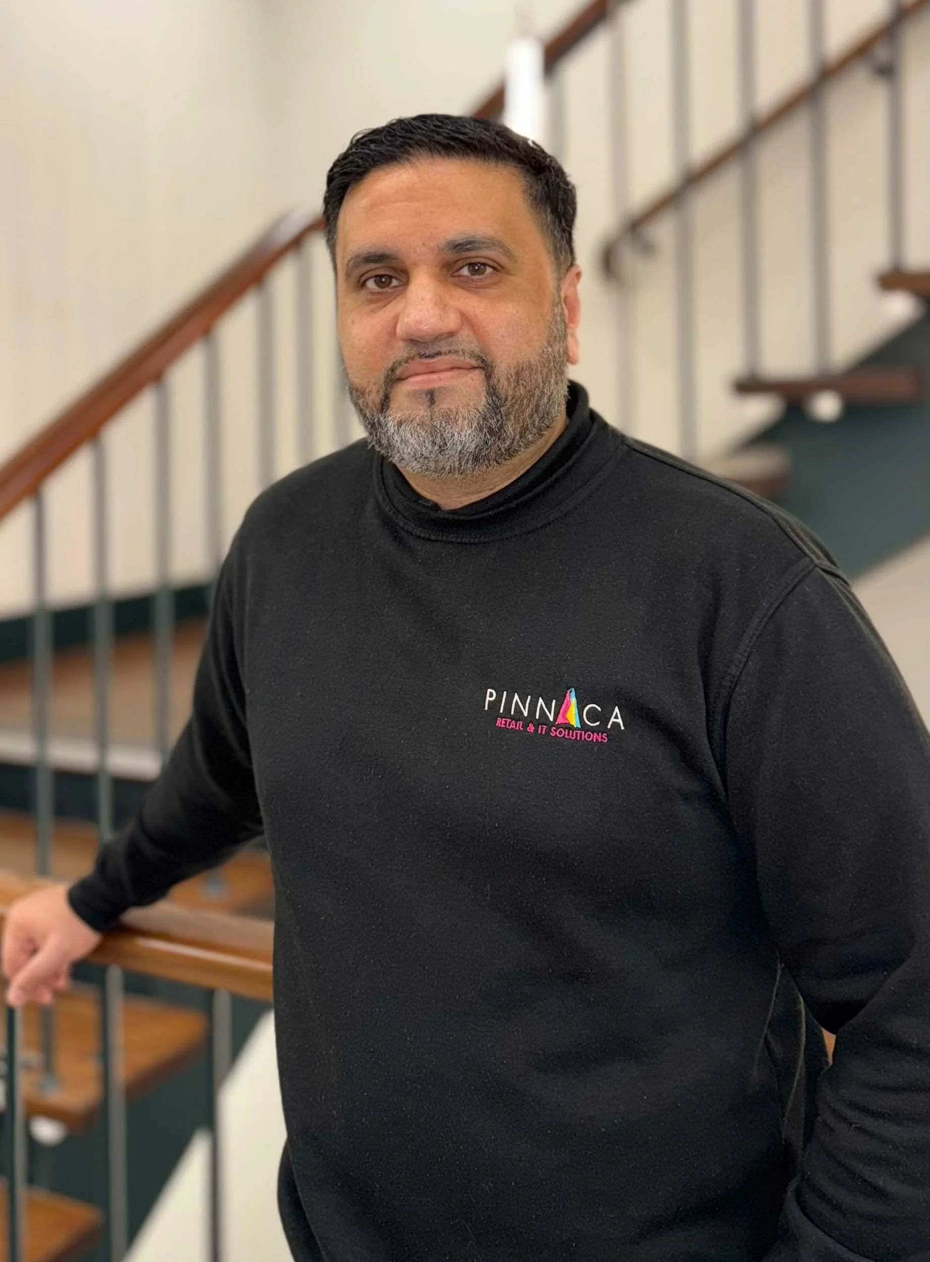 A man with short dark hair and a beard, wearing a black sweatshirt with a logo that says 'PINNA CA RETAIL & IT SOLUTIONS', standing inside a building with a staircase and a beige wall in the background.