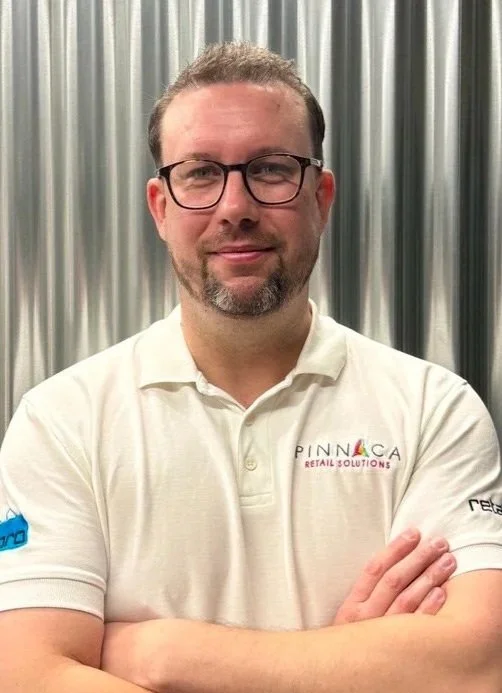 A smiling man with glasses and short brown hair, wearing a black polo shirt with a Pinnaca logo, standing in front of a staircase.