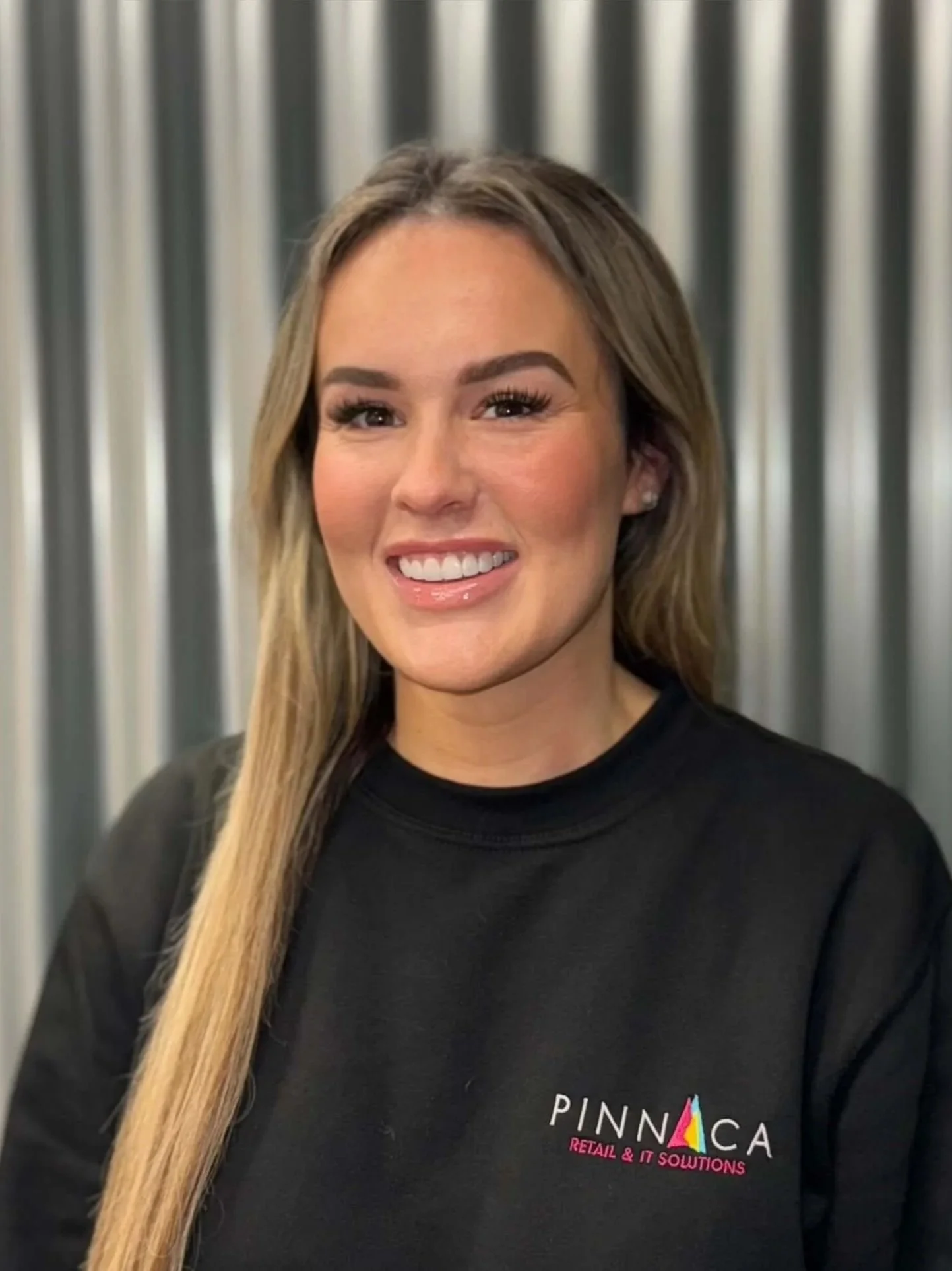 A woman smiling, wearing a black sweatshirt with the logo "PINNACA Retail & IT Solutions" and standing in front of a metallic, corrugated background.