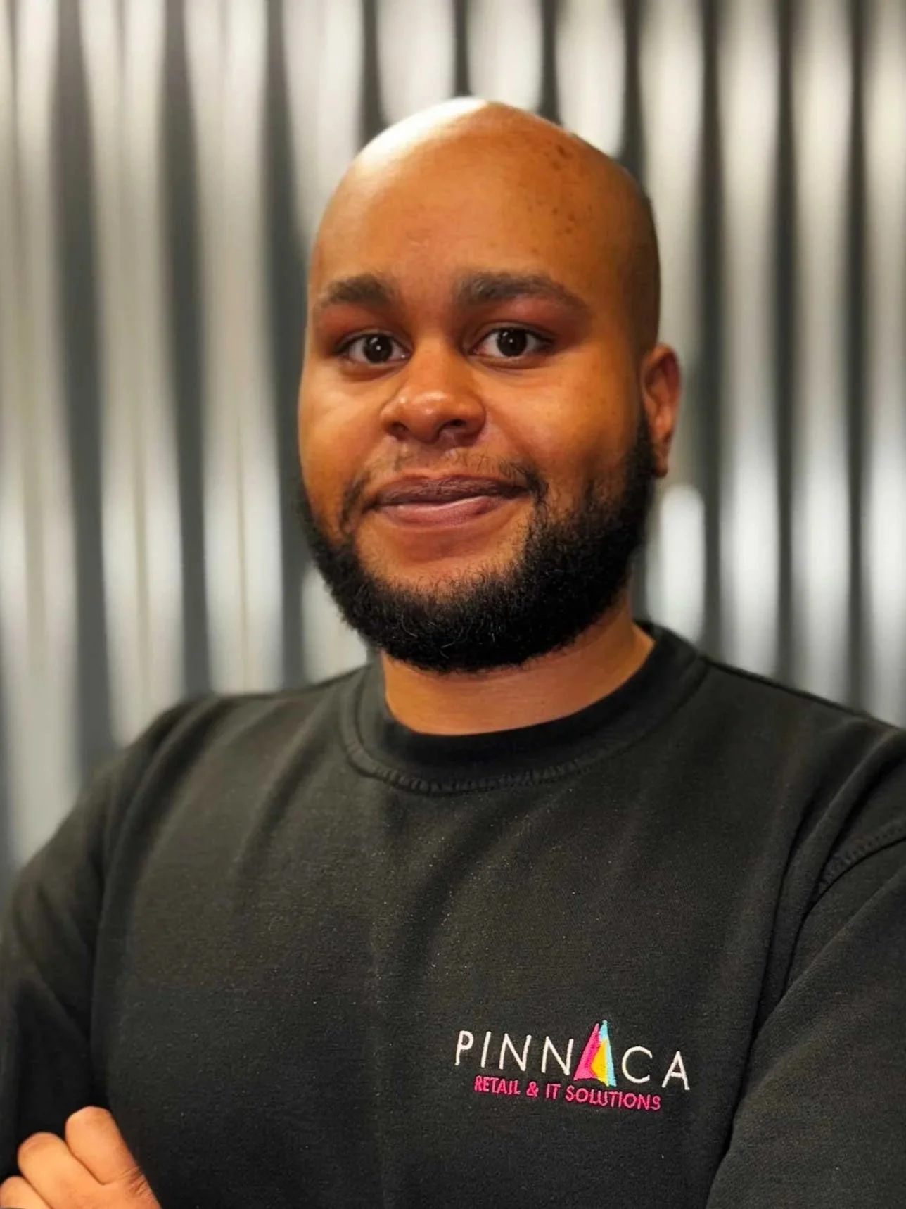 A man with a bald head and beard wearing a black sweatshirt with 'PINNACA RETAIL & IT SOLUTIONS' logo, standing with arms crossed, in front of a metallic striped background.