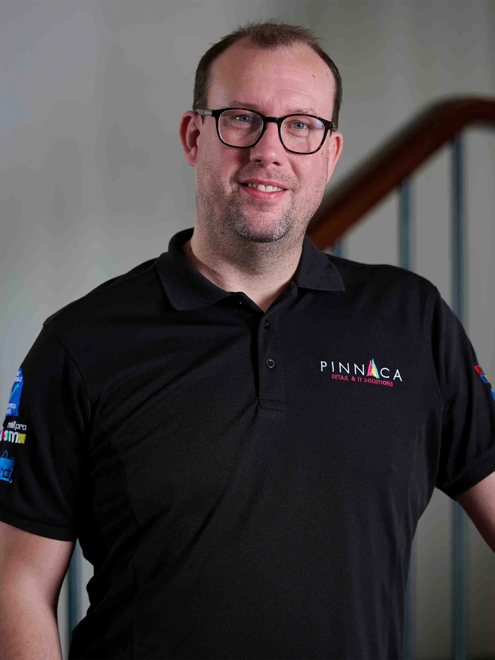 A smiling man with glasses and short brown hair, wearing a black polo shirt with a Pinnaca logo, standing in front of a staircase.