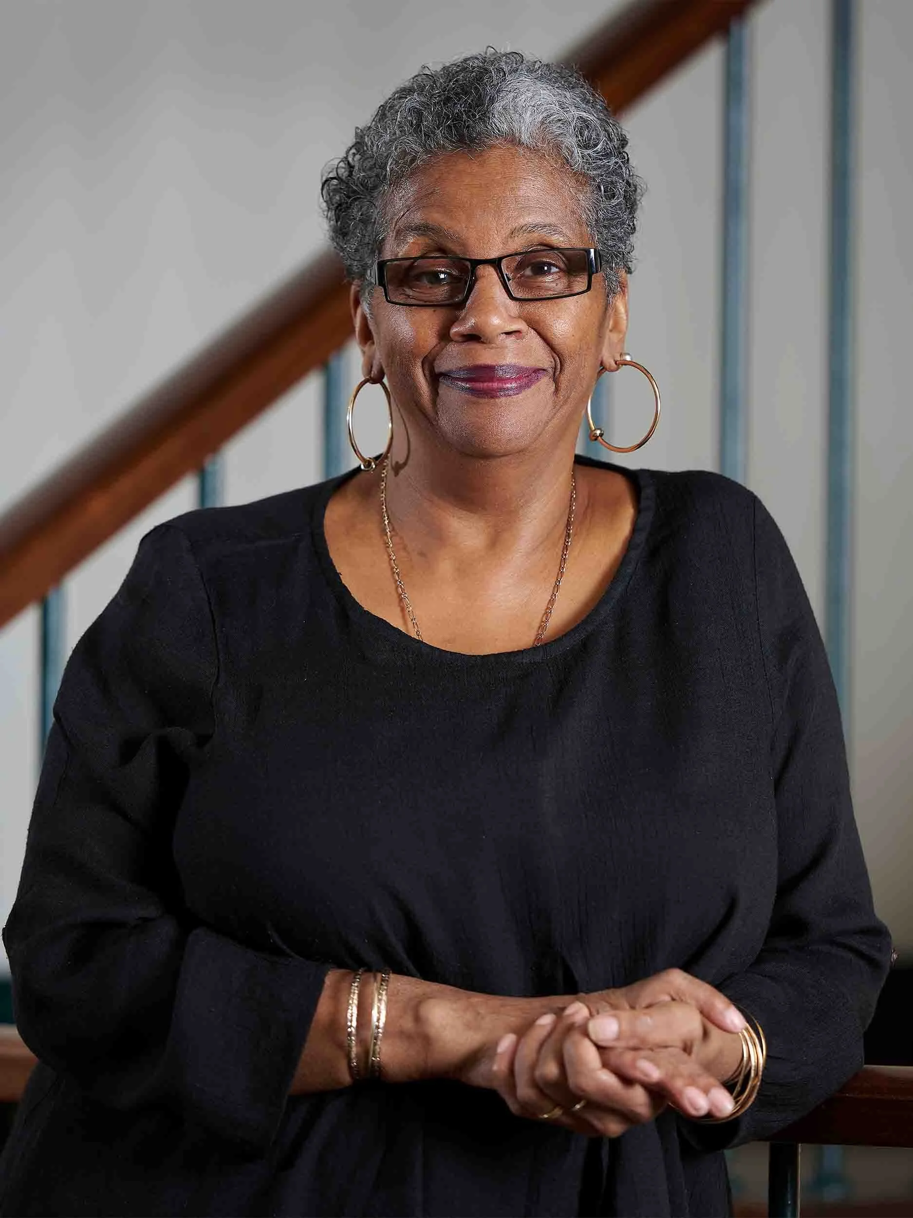 Portrait of an older woman with short curly gray hair, wearing glasses and jewelry, smiling in front of a staircase.
