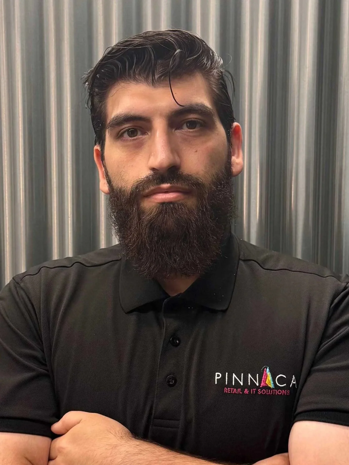 A man with dark hair and a full beard standing with arms crossed in front of a metallic background, wearing a black polo shirt with a Pinnaca logo.