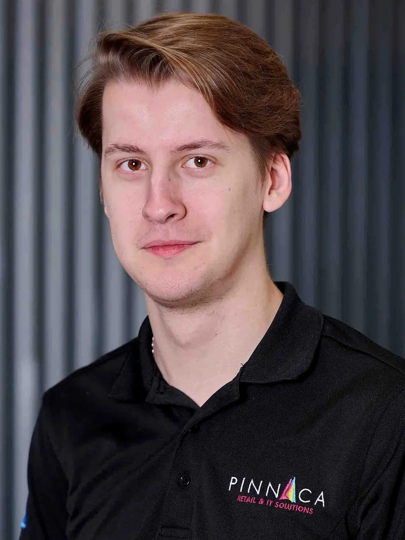 Young man with light skin, light brown hair, wearing a black polo shirt with logos, standing in front of a grey striped background.