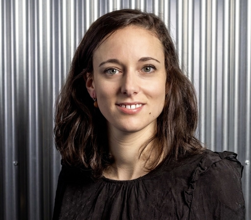 A woman with long brown hair and fair skin is standing indoors, smiling, wearing a black polo shirt with multiple sponsor logos."