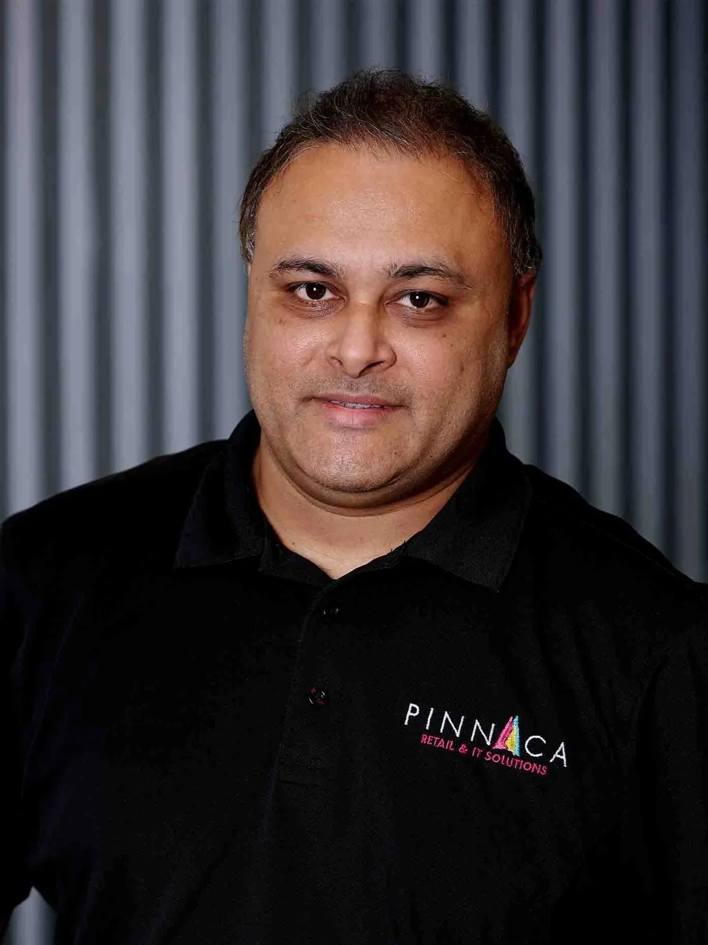 A man wearing a black polo shirt with logos stands in front of a metallic background.