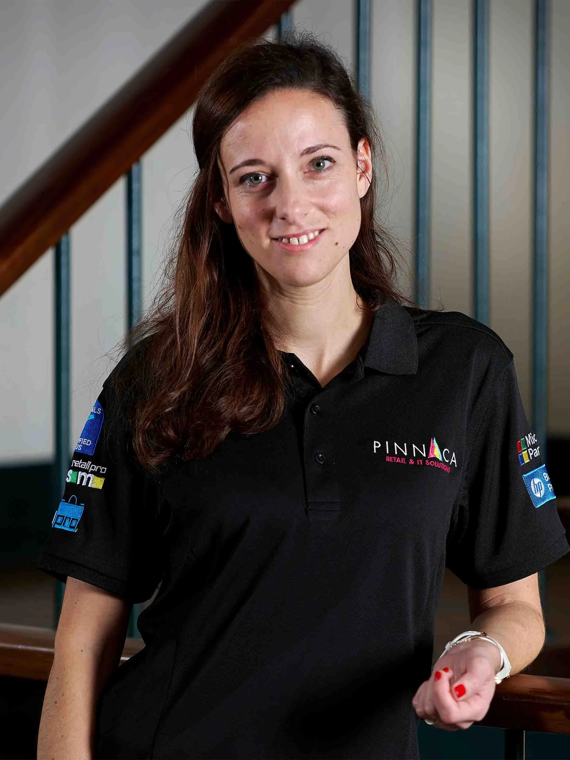 A woman with long brown hair and fair skin is standing indoors, smiling, wearing a black polo shirt with multiple sponsor logos."